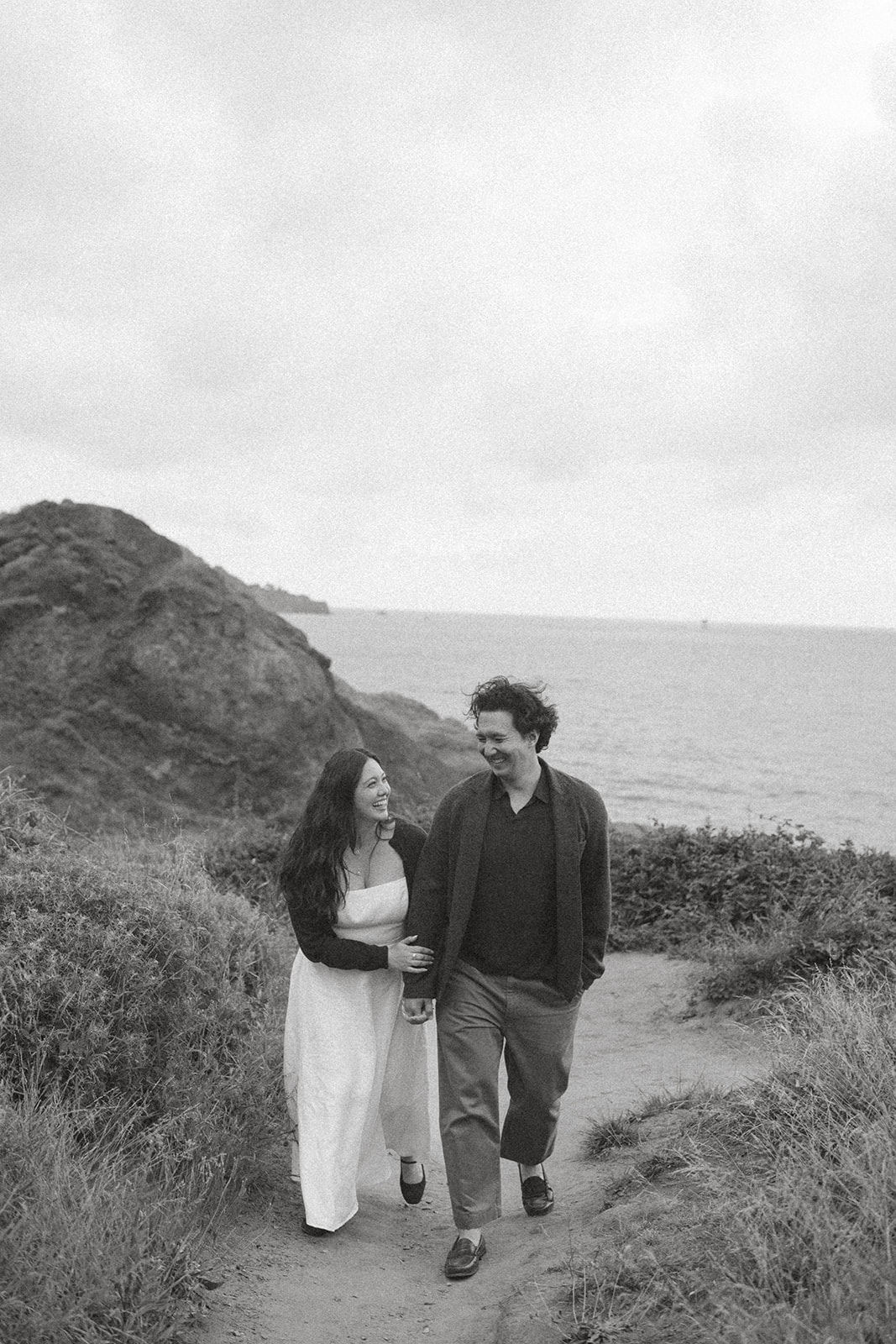 Black-and-white photo of a couple walking hand in hand along a coastal path, smiling at each other with the ocean and rugged cliffs behind them.