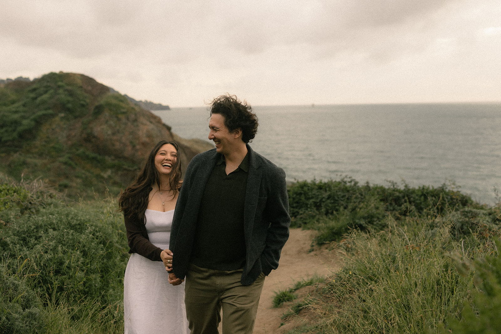 Couple walking hand in hand along a cliffside trail overlooking the ocean, framed by coastal greenery under an overcast sky.