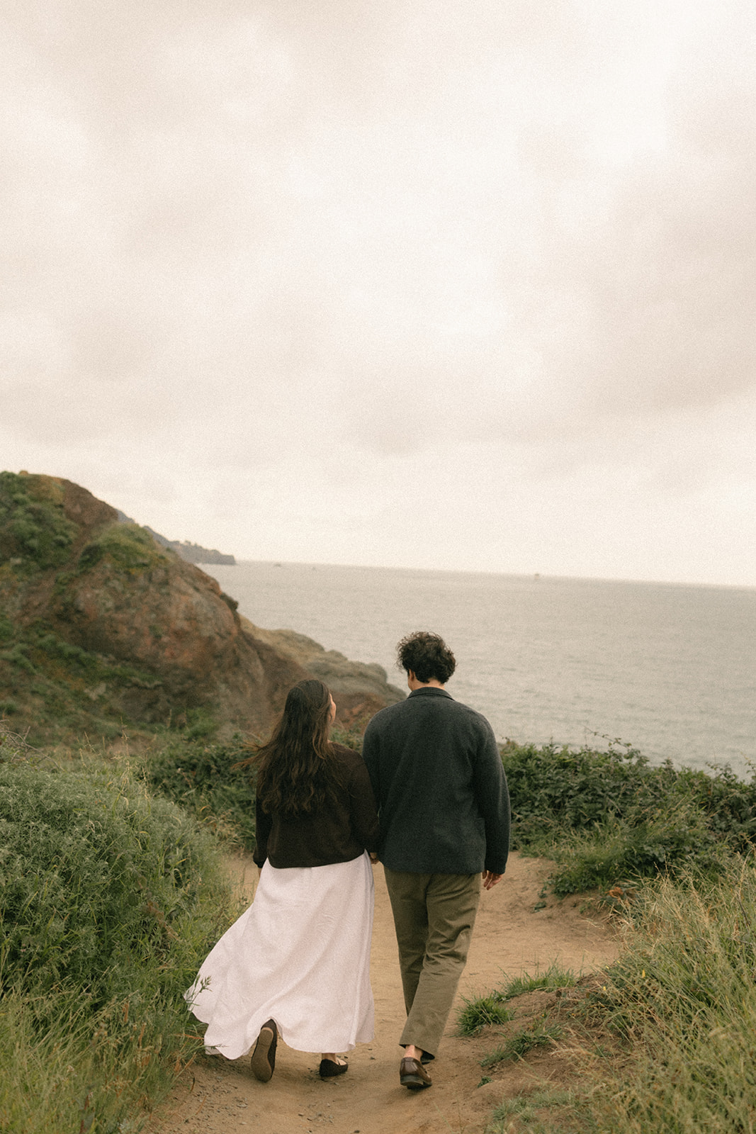 Couple walking away together on the Battery to Bluffs Trail, surrounded by coastal greenery with rocky cliffs and the Pacific Ocean stretching into the distance behind them.