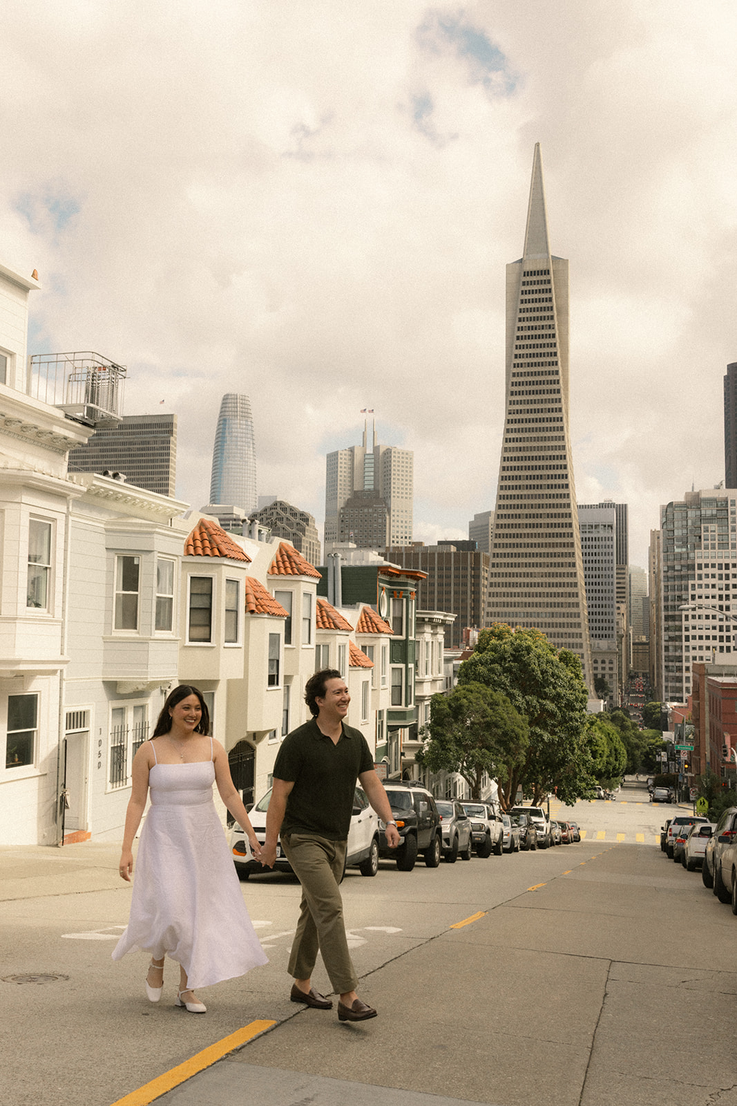 Couple holding hands while walking down a steep hill with the Transamerica Pyramid, downtown skyline, and historic homes lining the San Francisco City Streets during a relaxed San Francisco engagement photos session.