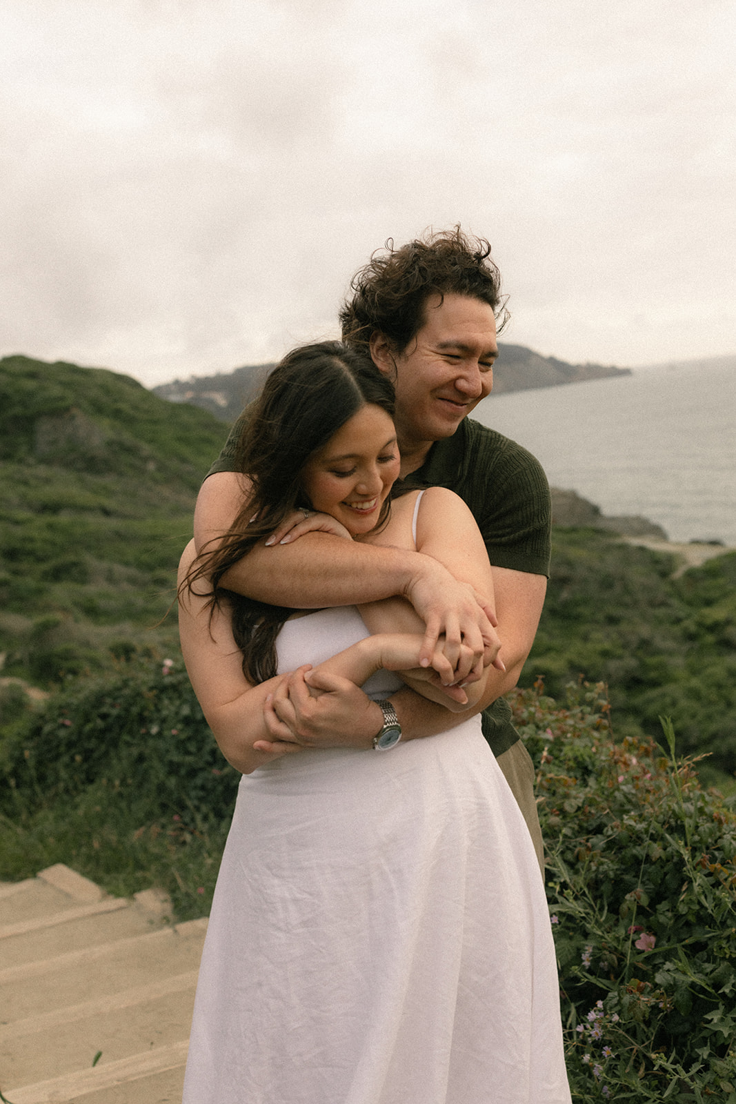 Couple embracing on the Battery to Bluffs Trail, surrounded by coastal greenery with ocean cliffs and the Pacific shoreline stretching into the background.