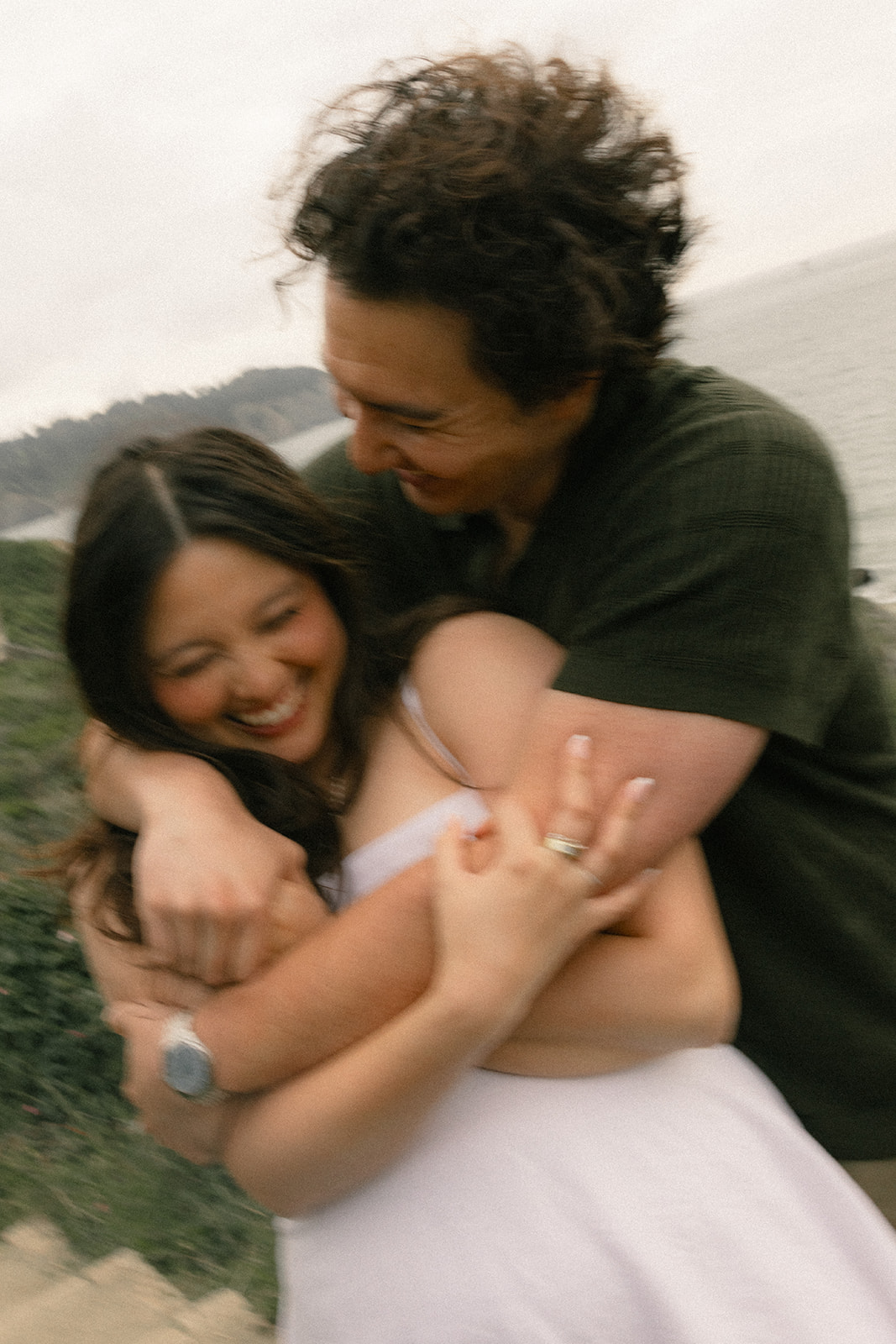 Close-up of a couple laughing as one embraces the other from behind on a coastal overlook, with the ocean softly blurred in the background.