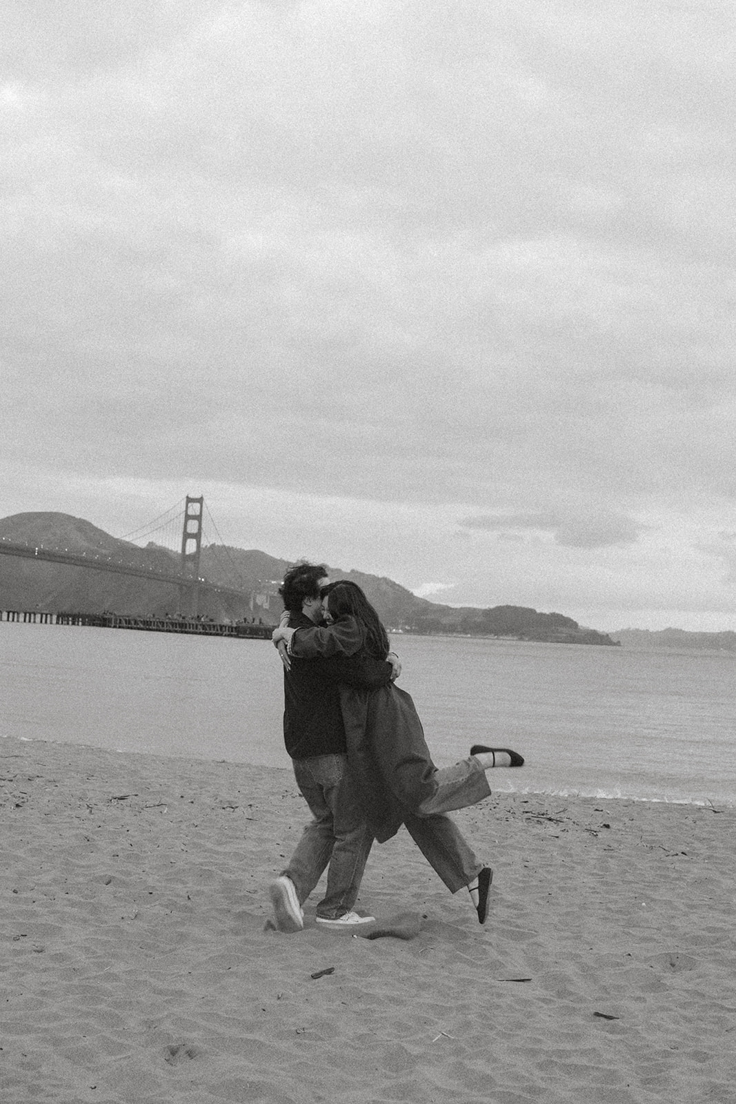 Black-and-white photo of a couple embracing on a sandy beach with the Golden Gate Bridge in the background, one partner lifting a foot in a playful hug for engagement photos by San Francisco photographer