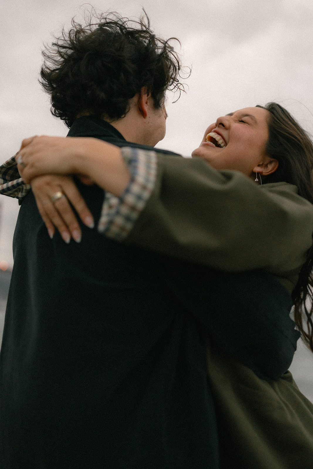 Close-up of a couple embracing and laughing outdoors near the waterfront, captured from a low angle against a cloudy sky.