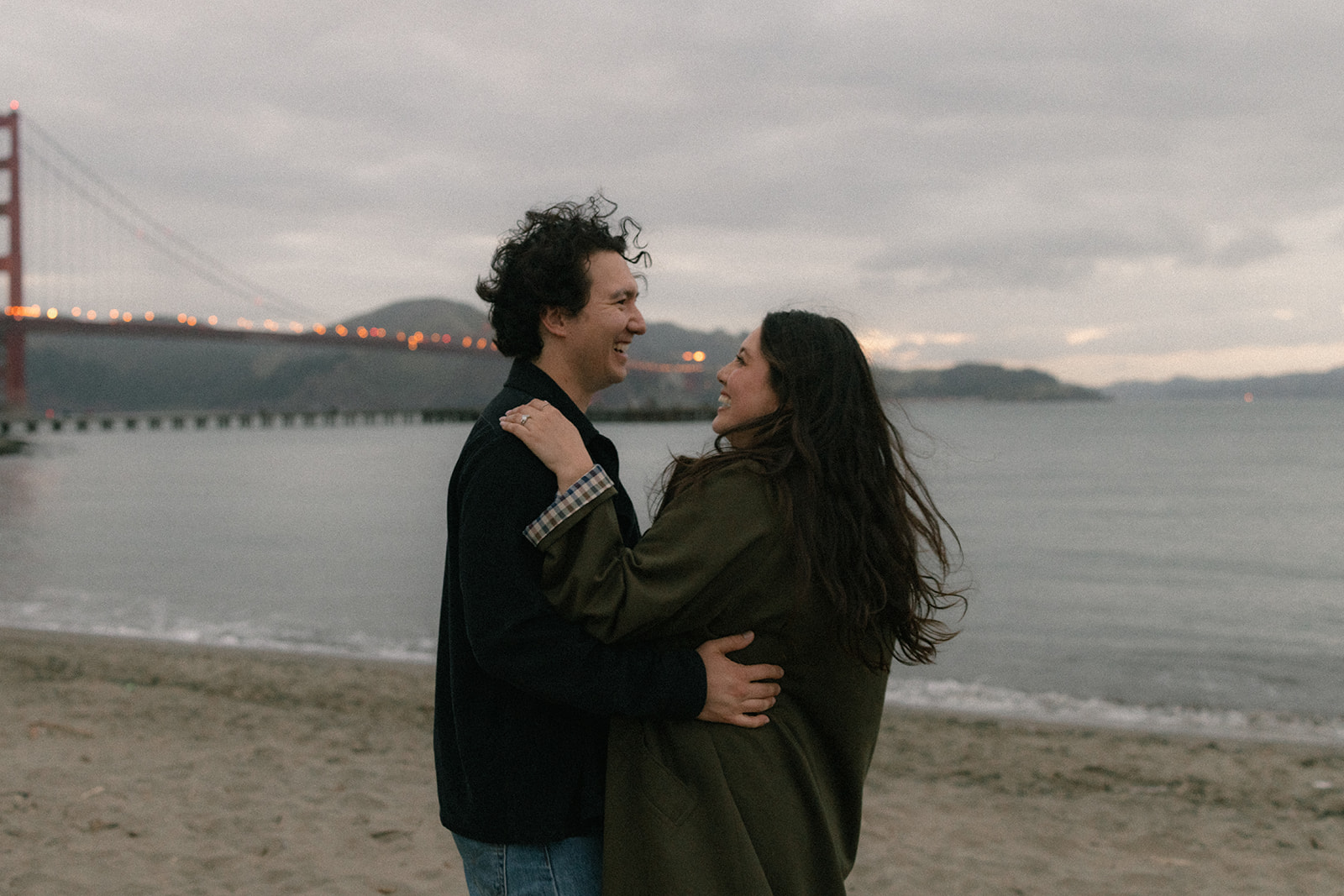 Couple embracing on the beach at sunset with the Golden Gate Bridge and calm waterfront in the background.