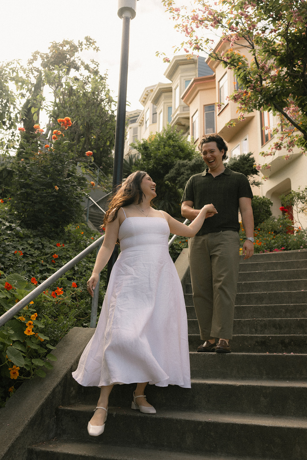 Laughing couple walking down a flower-lined neighborhood staircase surrounded by lush gardens, blooming trees, and colorful homes, photographed by a San Francisco photographer.