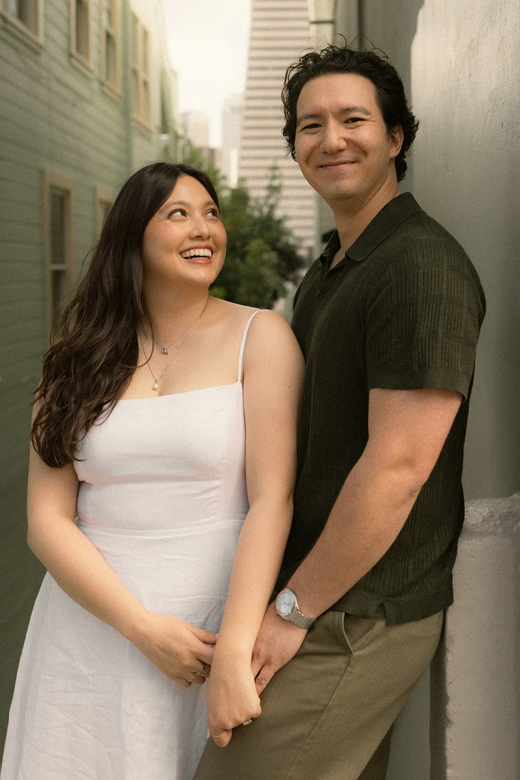 Smiling couple holding hands in a narrow alley with the Transamerica Pyramid rising between colorful buildings in the background during a San Francisco engagement photos session.