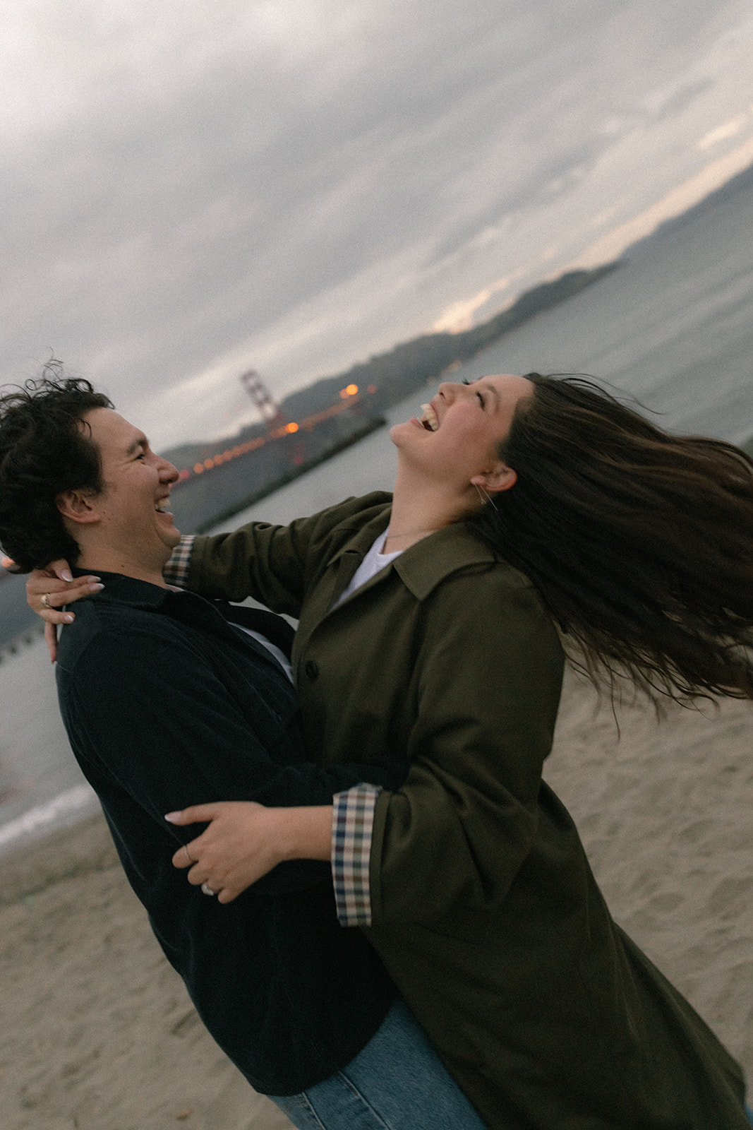 Couple laughing while spinning together on the beach at dusk, with the Golden Gate Bridge softly visible behind them.