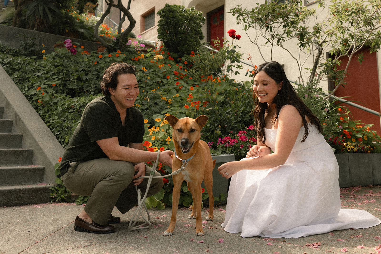 Couple kneeling beside their dog on a flower-lined residential sidewalk, with colorful gardens, stairways, and charming homes creating a vibrant neighborhood scene photographed by a San Francisco photographer.