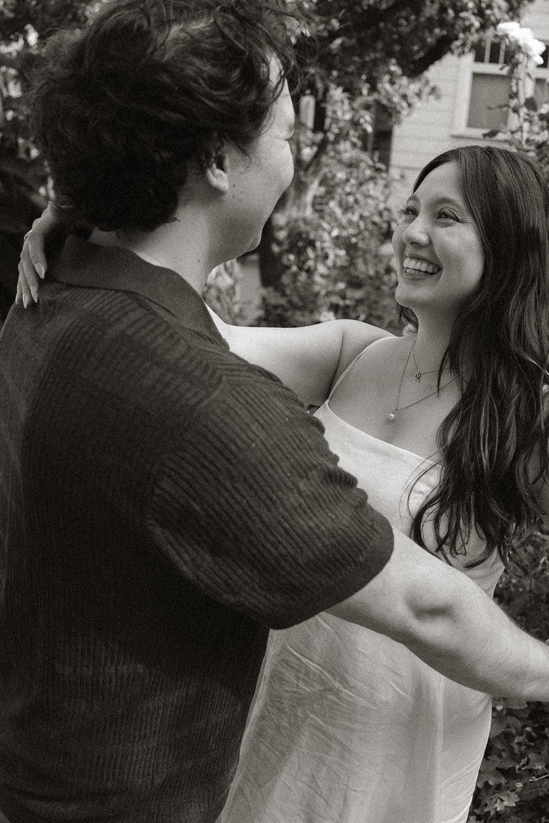 Black and white portrait of a smiling couple embracing in a lush neighborhood garden, with blooming flowers and leafy trees creating a romantic backdrop during a San Francisco engagement photos session.