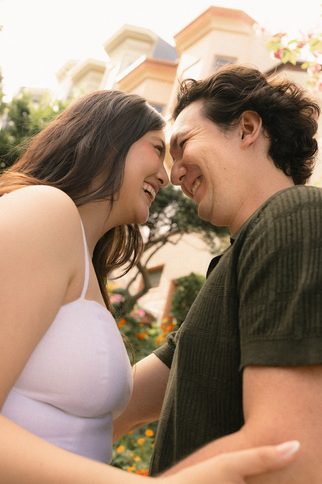 Close-up portrait of a smiling couple leaning forehead to forehead, with blooming flowers, lush greenery, and hillside homes softly blurred in the background during a San Francisco engagement photos session.