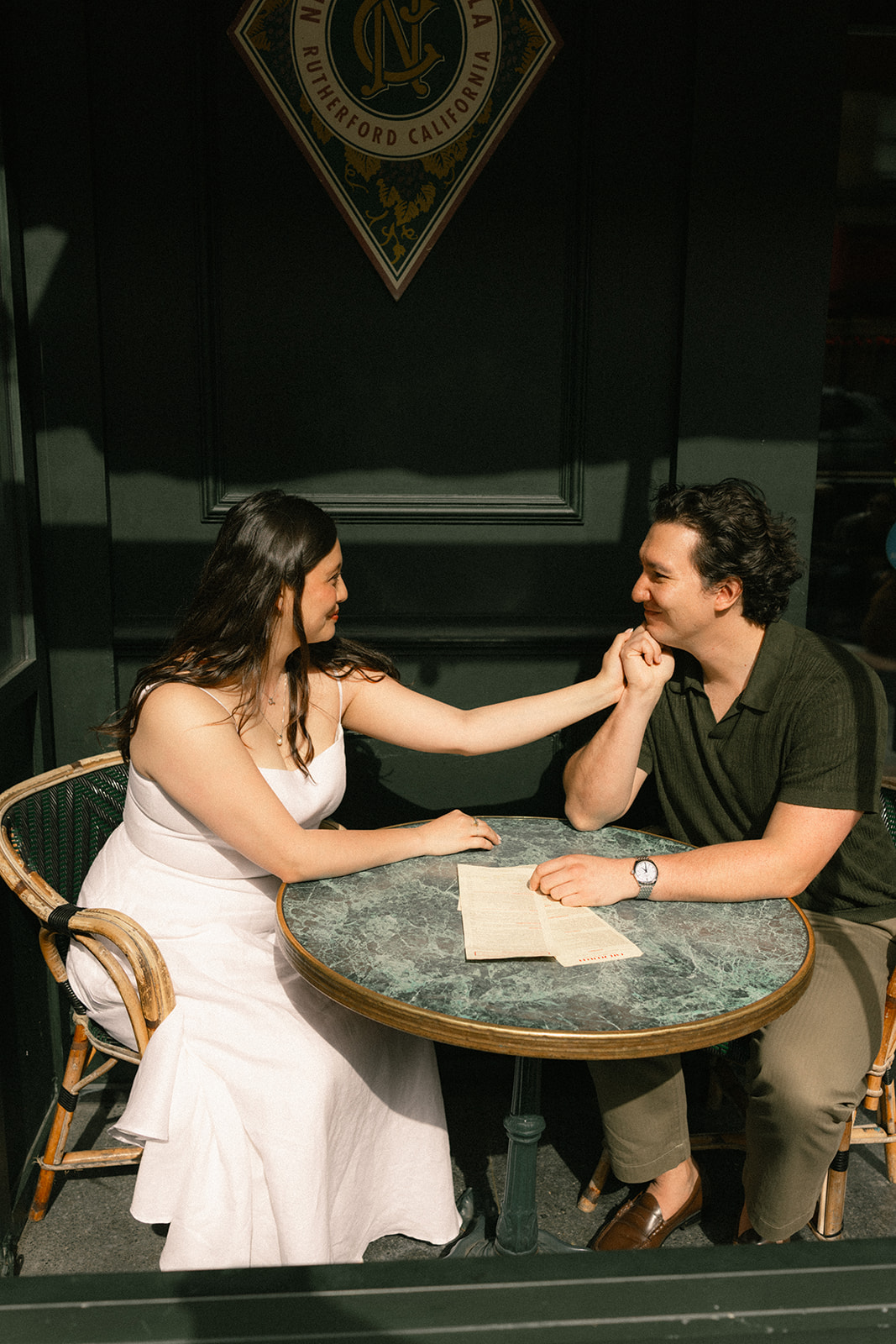 Couple sitting together at an outdoor café table, smiling and holding hands in front of a dark storefront, photographed by a San Francisco photographer.
