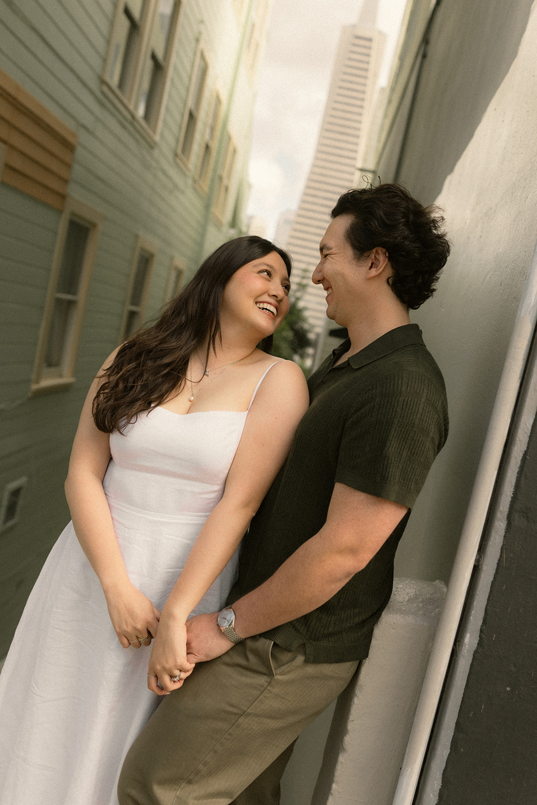 Couple smiling at each other while standing in a narrow alley framed by pastel buildings, with the Transamerica Pyramid rising in the background during a San Francisco engagement photos session.