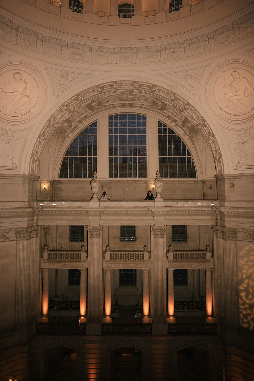 Wide architectural San Francisco City Hall wedding photo showing the couple beneath the grand rotunda, ornate arches, columns, and warm evening lighting.