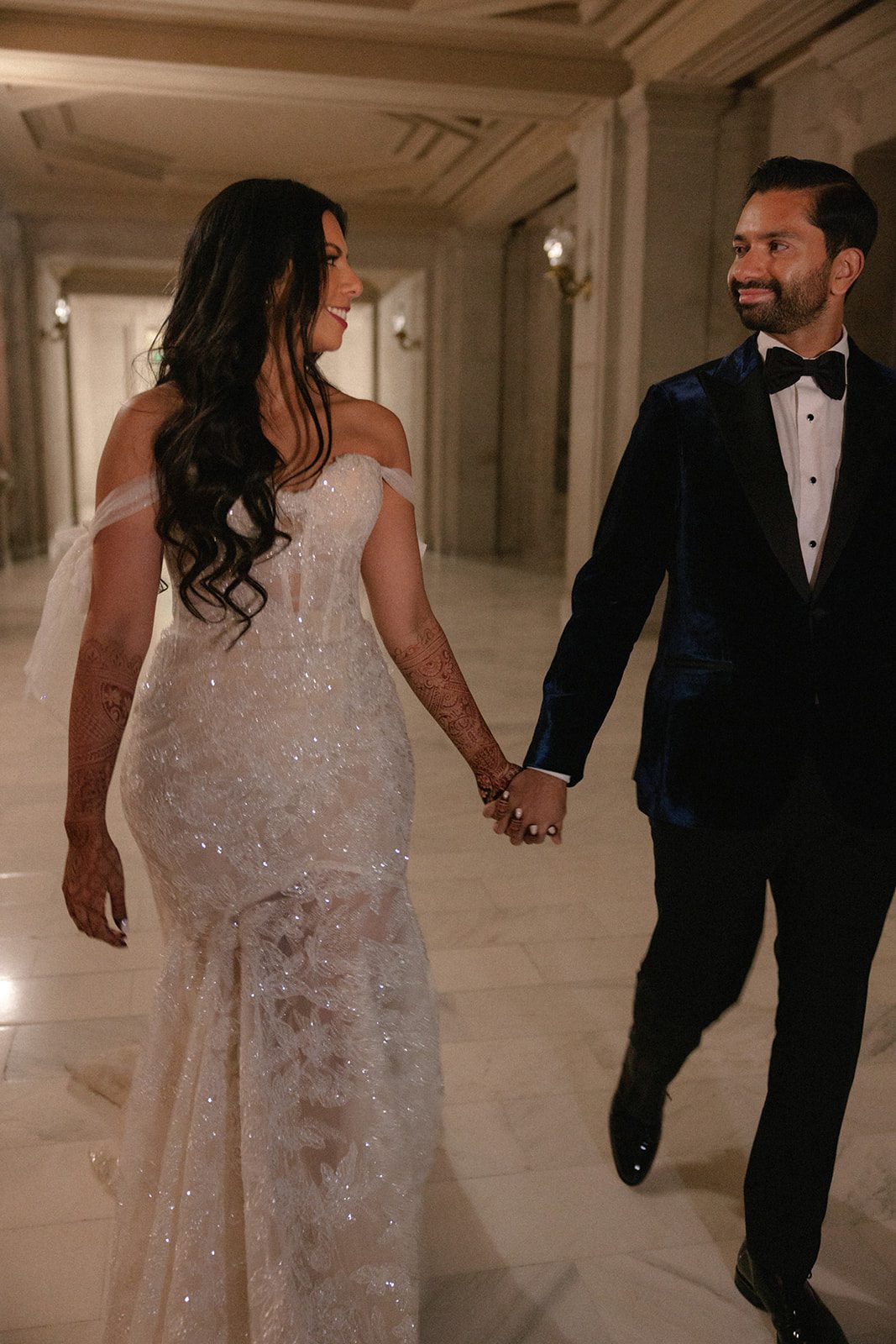 Bride and groom walk hand in hand through a marble hallway for candid San Francisco City Hall wedding photos, wearing a sparkling gown and velvet tuxedo.