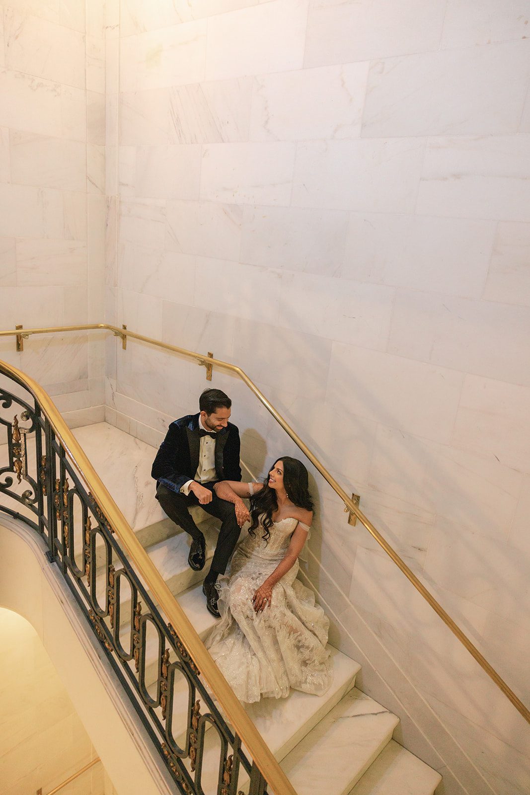 Newlyweds sit together on a marble staircase during their portrait session, captured by a San Francisco City Hall photographer beside ornate iron and gold railings.