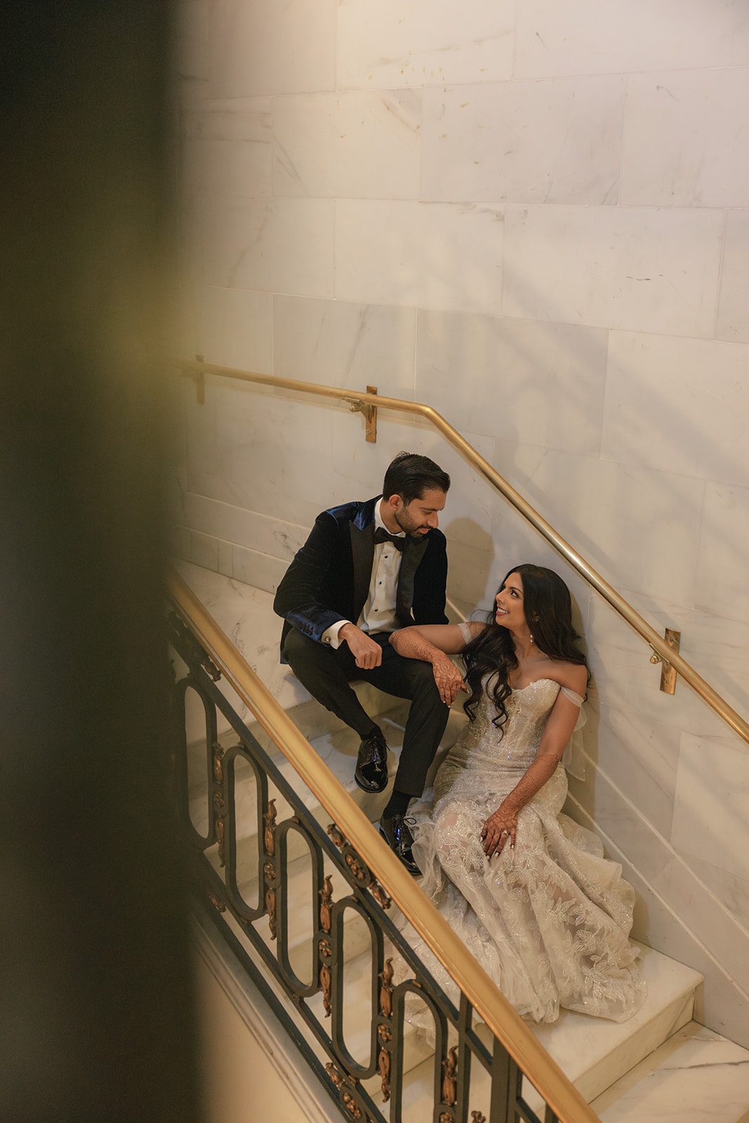 Bride and groom sit together on a marble staircase during their portrait session, captured by a San Francisco City Hall photographer beside ornate black-and-gold railings.