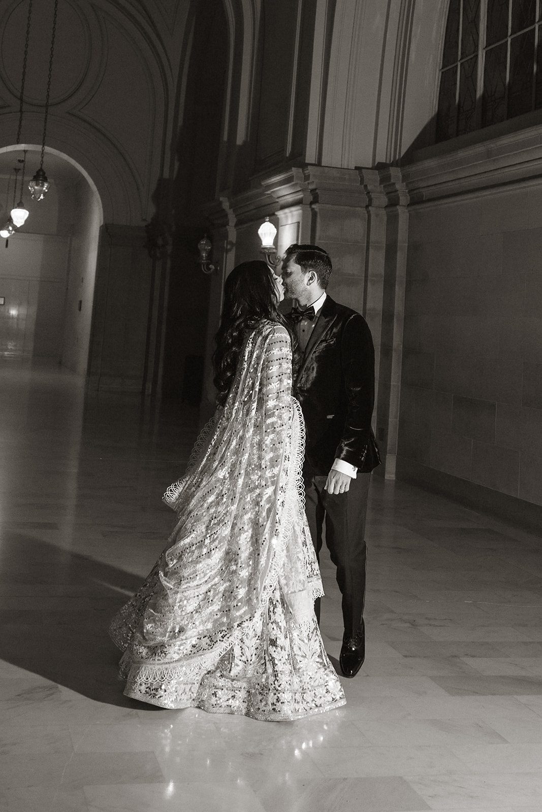 Black-and-white San Francisco City Hall wedding portrait of a couple kissing beneath dramatic arched hallways and vintage lights.