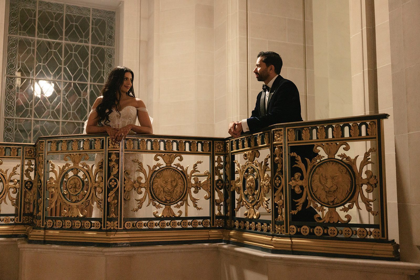 Bride and groom pose across an ornate gold railing for San Francisco City Hall wedding photos, framed by tall stone columns and decorative windows.