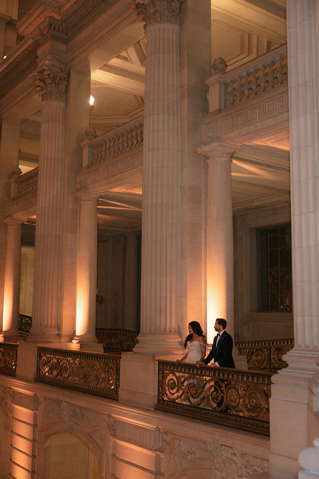 Newlyweds stand along an ornate balcony beneath towering columns during their San Francisco City Hall wedding, surrounded by warm architectural lighting.