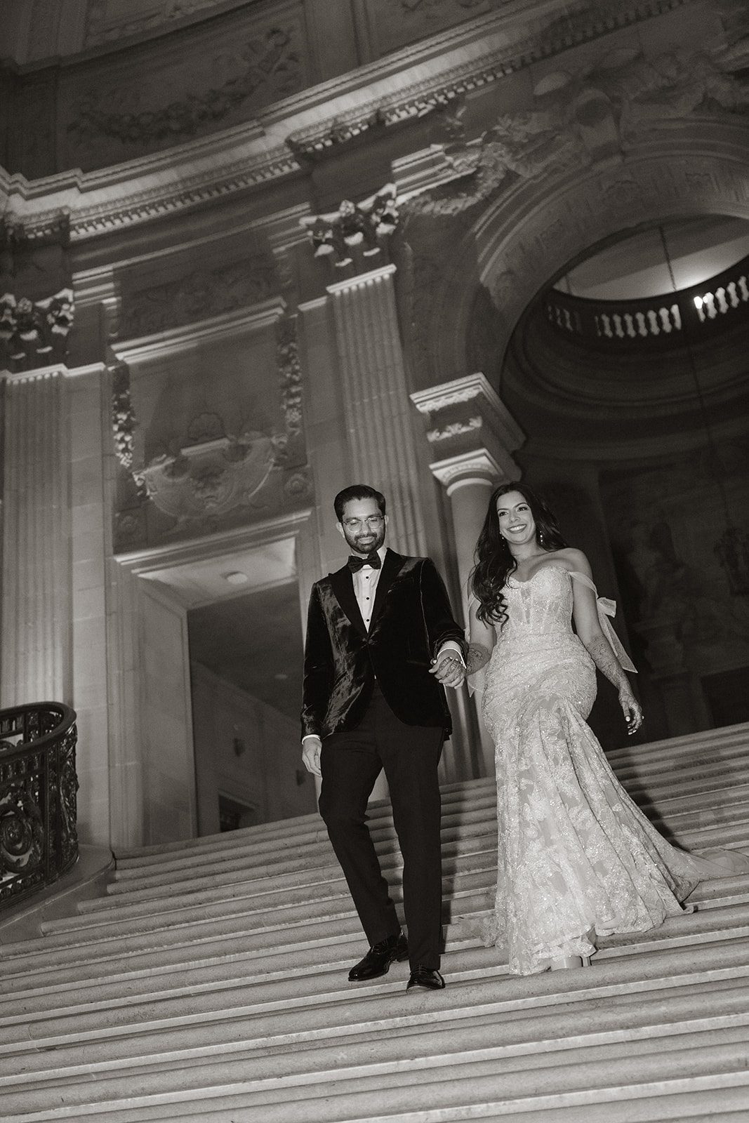 Black-and-white San Francisco City Hall wedding photos of the bride and groom walking hand in hand down the grand marble staircase beneath ornate arches.