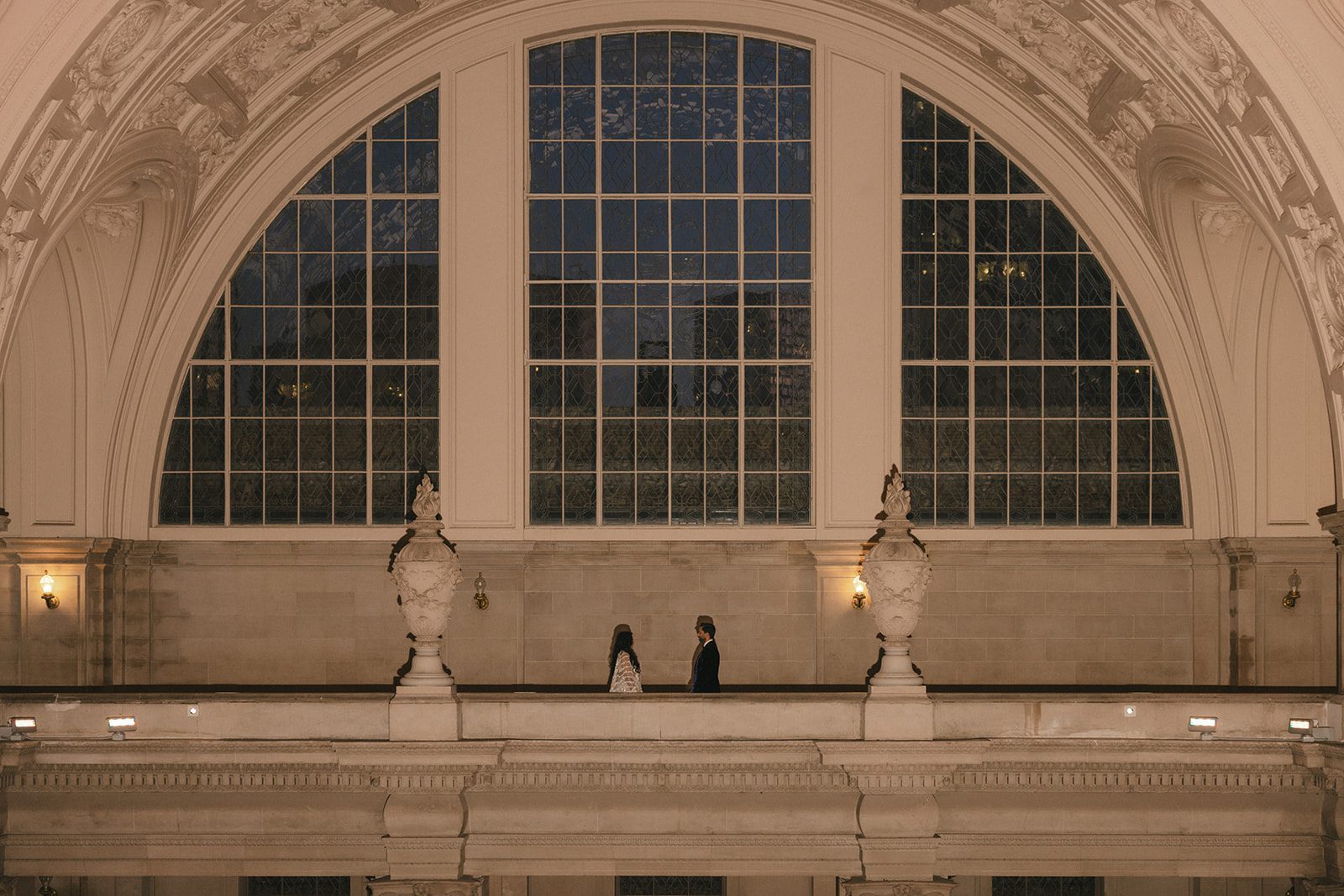 Wide San Francisco City Hall wedding portrait of the couple facing each other beneath a grand arched window and decorative architectural details.