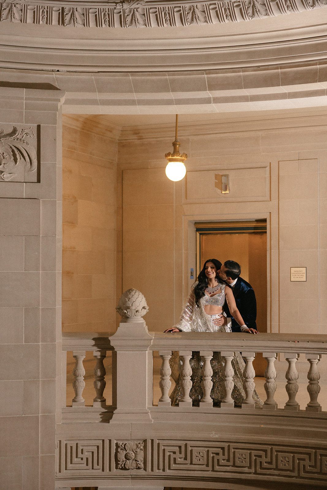 Groom kisses the bride on a balcony during their San Francisco City Hall wedding, framed by ornate stone architecture and warm lighting.