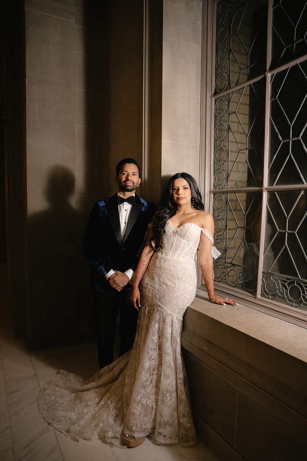 Bride and groom pose beside a decorative window for San Francisco City Hall wedding photos, highlighting her beaded gown, henna, and his velvet tuxedo.