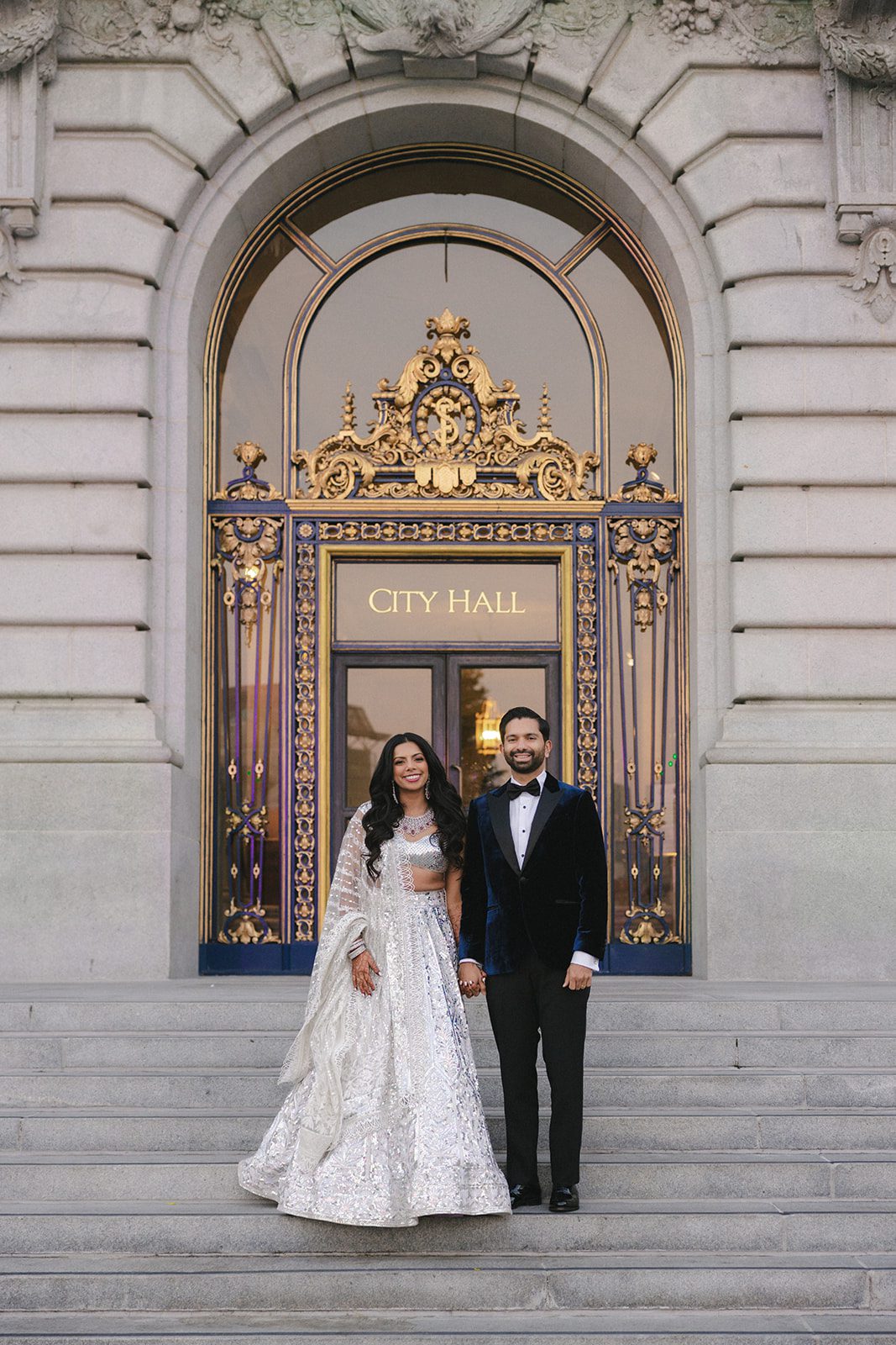 Newlyweds pose beneath the ornate gold-trimmed entrance for San Francisco City Hall wedding photos, with the bride in a shimmering lehenga and groom in a velvet tuxedo.