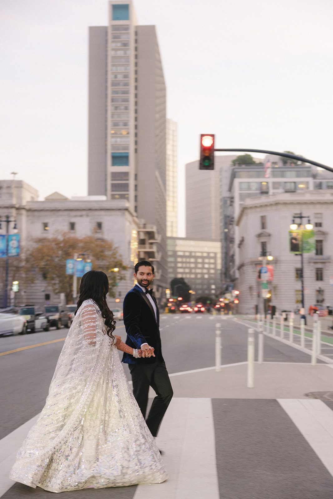 Newlyweds cross a downtown street hand in hand after their San Francisco City Hall wedding, with the bride’s shimmering lehenga and groom’s velvet tuxedo against the city skyline.
