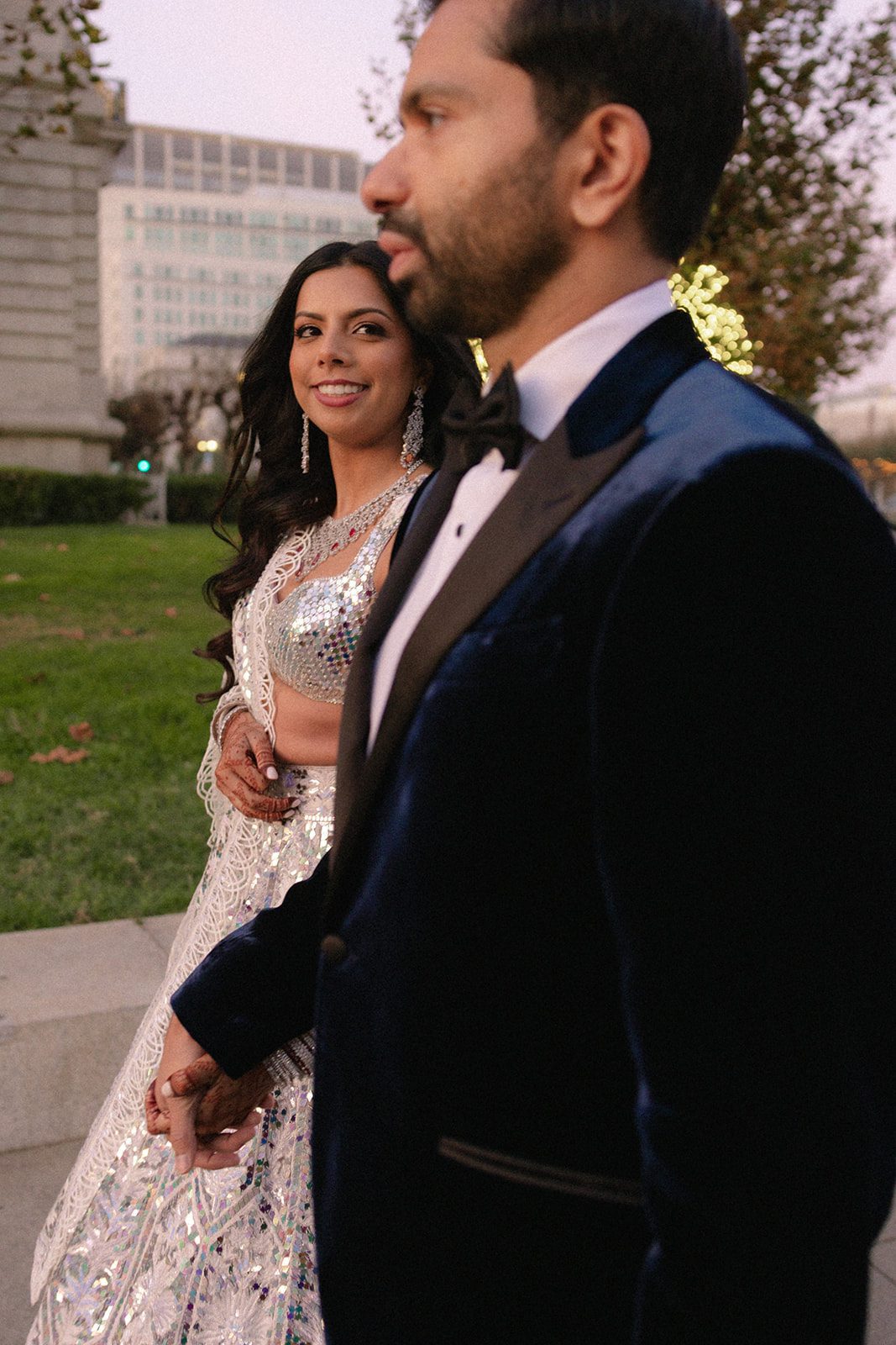 Candid portrait of the newlyweds walking outside City Hall, captured by a Bay area wedding photographer with the bride smiling toward the groom at dusk.