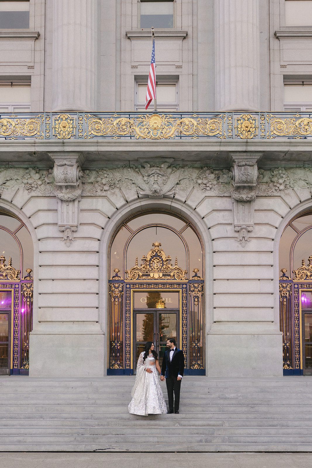 Couple poses outside the landmark entrance after their San Francisco City Hall wedding, framed by the grand stone façade, gilded doors, and American flag.