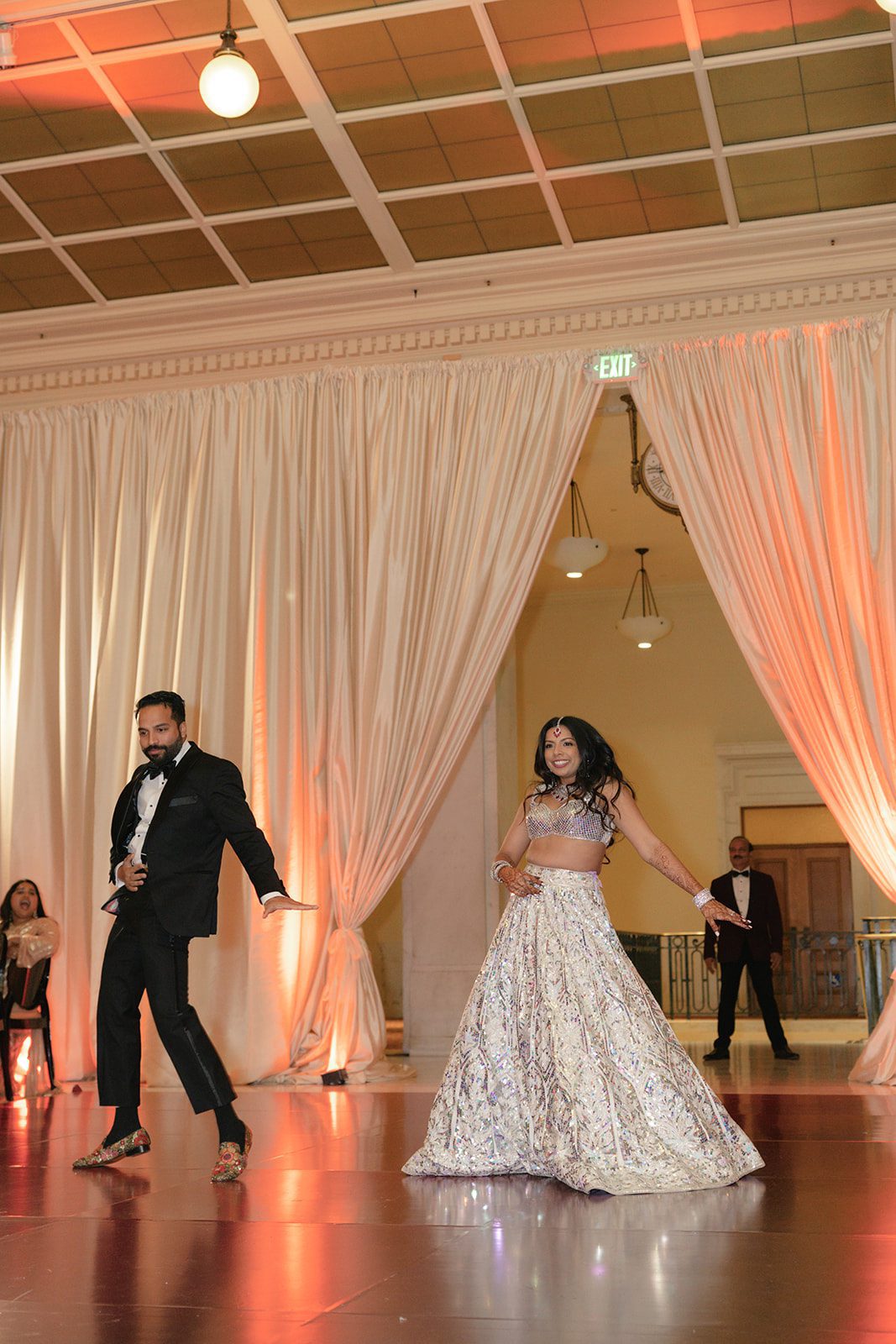 Bride and groom dance together during their San Francisco City Hall wedding reception, with her shimmering lehenga and his velvet tuxedo illuminated by warm reception lighting.