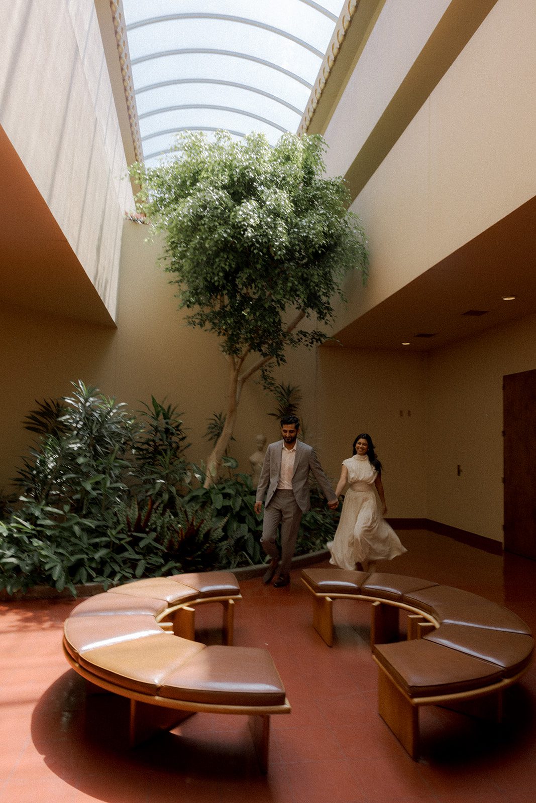 Couple holding hands while walking through the indoor garden atrium during their Marin County Civic CenterElopement, framed by a tall tree, lush plants, curved seating, and skylight, photographed by a Bay Area wedding photographer.
