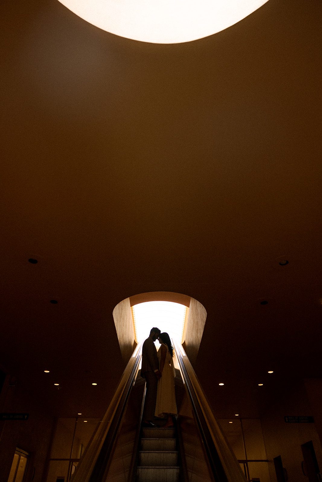 Silhouette of a couple touching foreheads on an escalator during their Marin County Civic CenterElopement, framed by a glowing skylight and dramatic interior architecture, captured by a Bay Area wedding photographer.