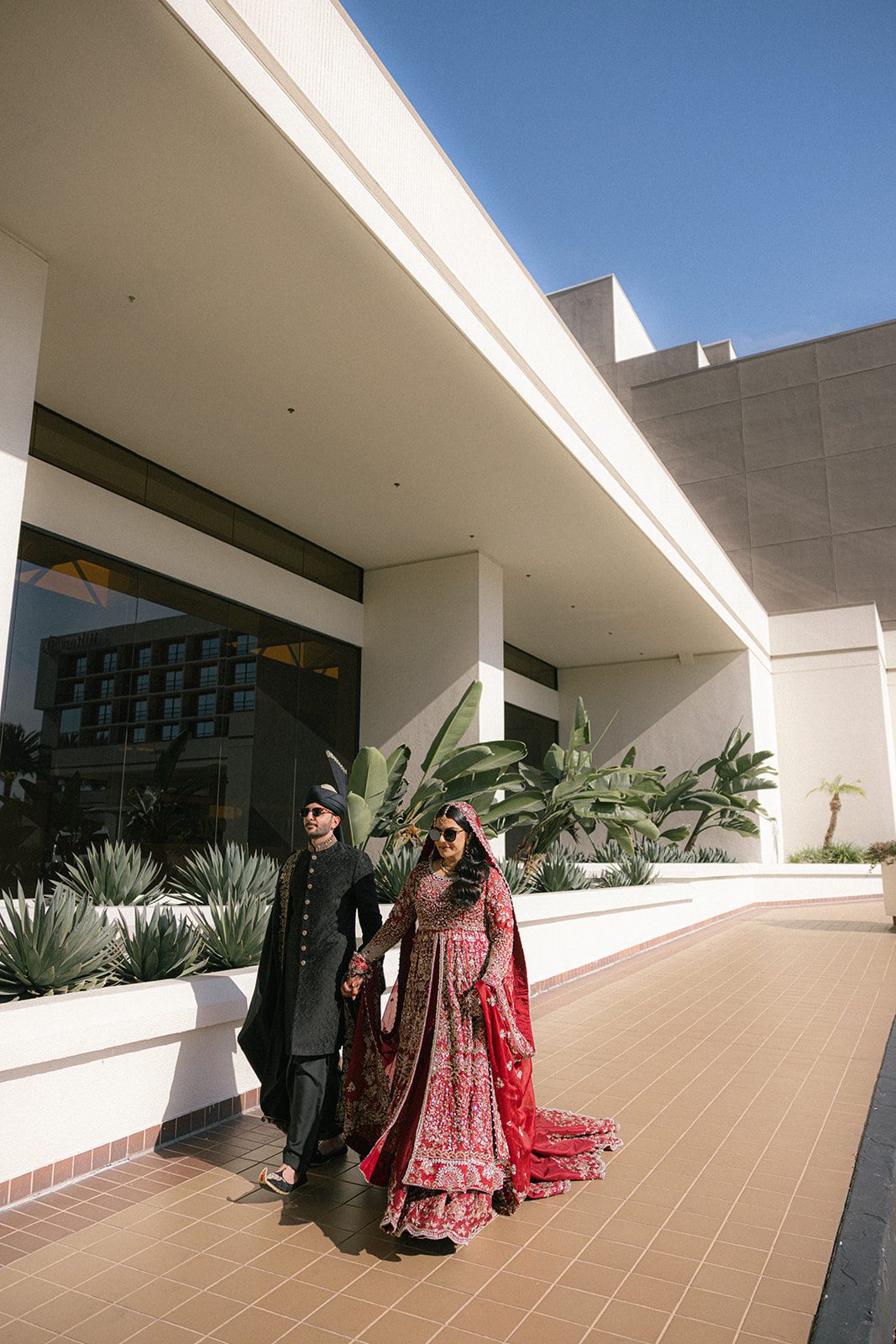 Bride in a red embroidered lehenga and groom in black traditional attire walking hand in hand outdoors, captured by an indian wedding photographer during their wedding portraits.