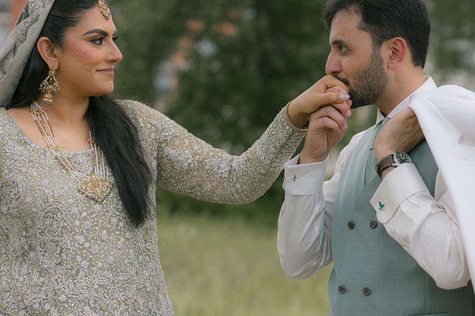 Groom kissing the bride’s hand during an outdoor south asian wedding portrait, highlighting her silver embroidered attire and traditional jewelry.