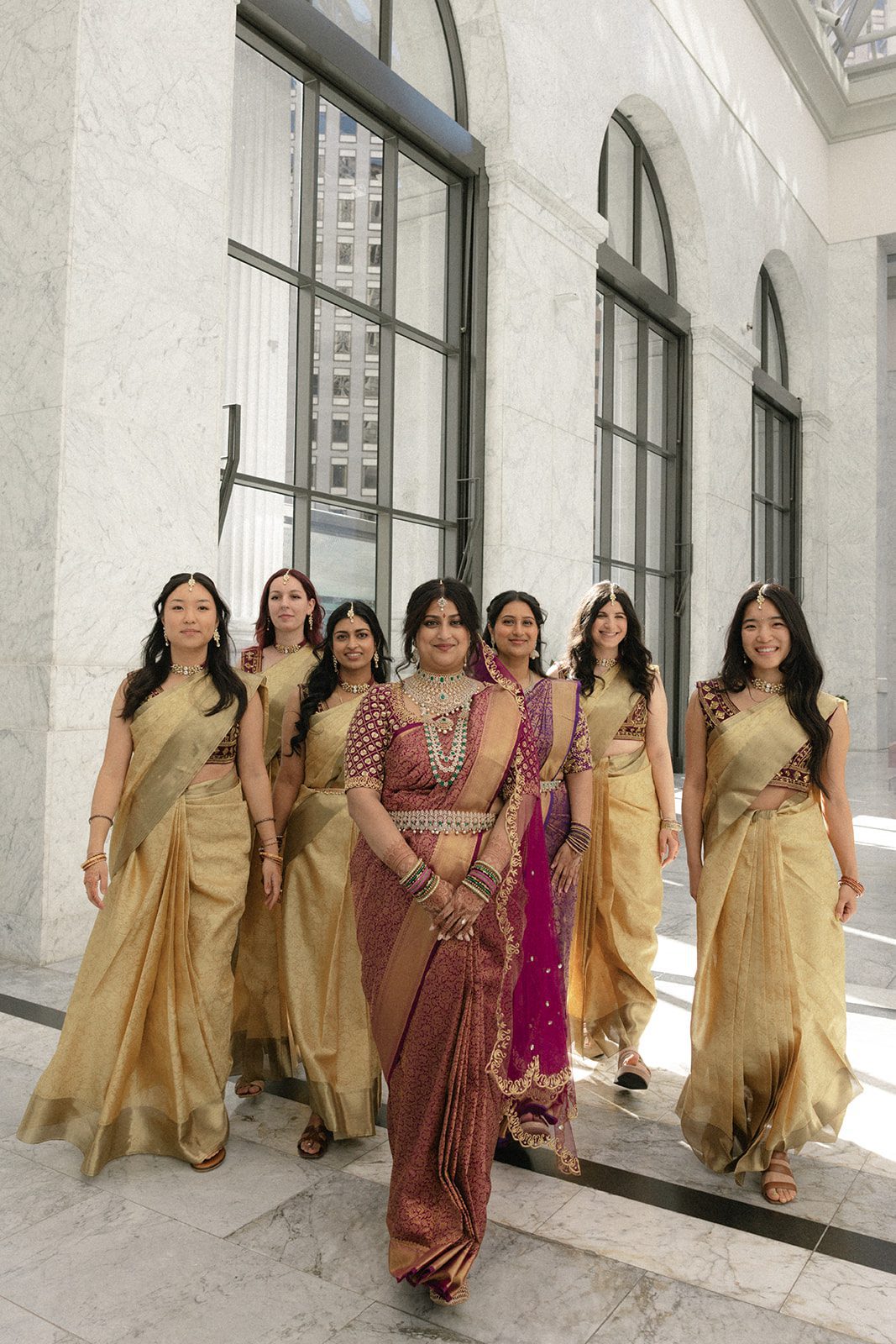 Bride walking with her wedding party in gold saris through the marble interior of the Conservatory at One Sansome.
