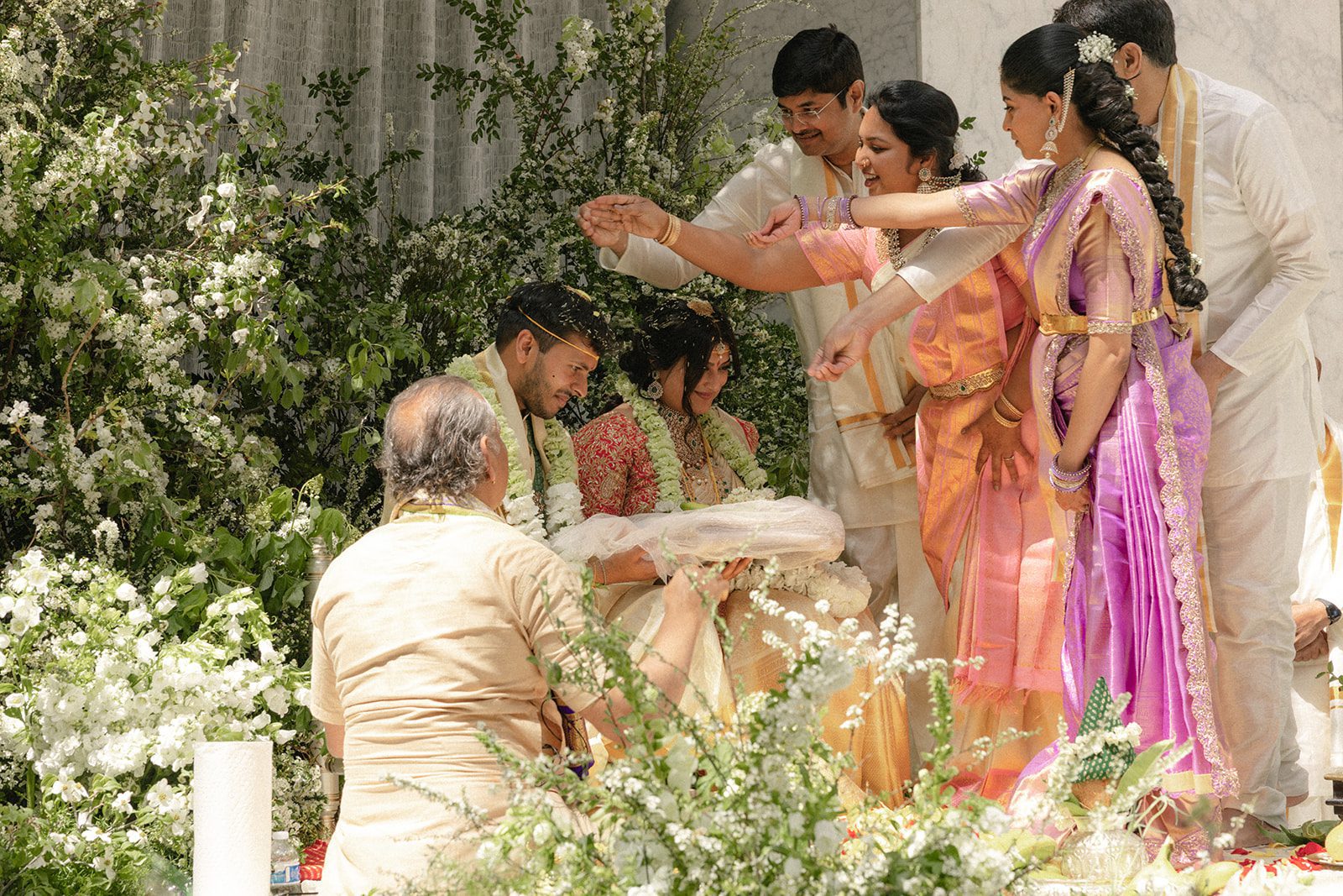 Family members participate in a traditional Telugu wedding ritual with the bride and groom surrounded by abundant white florals.