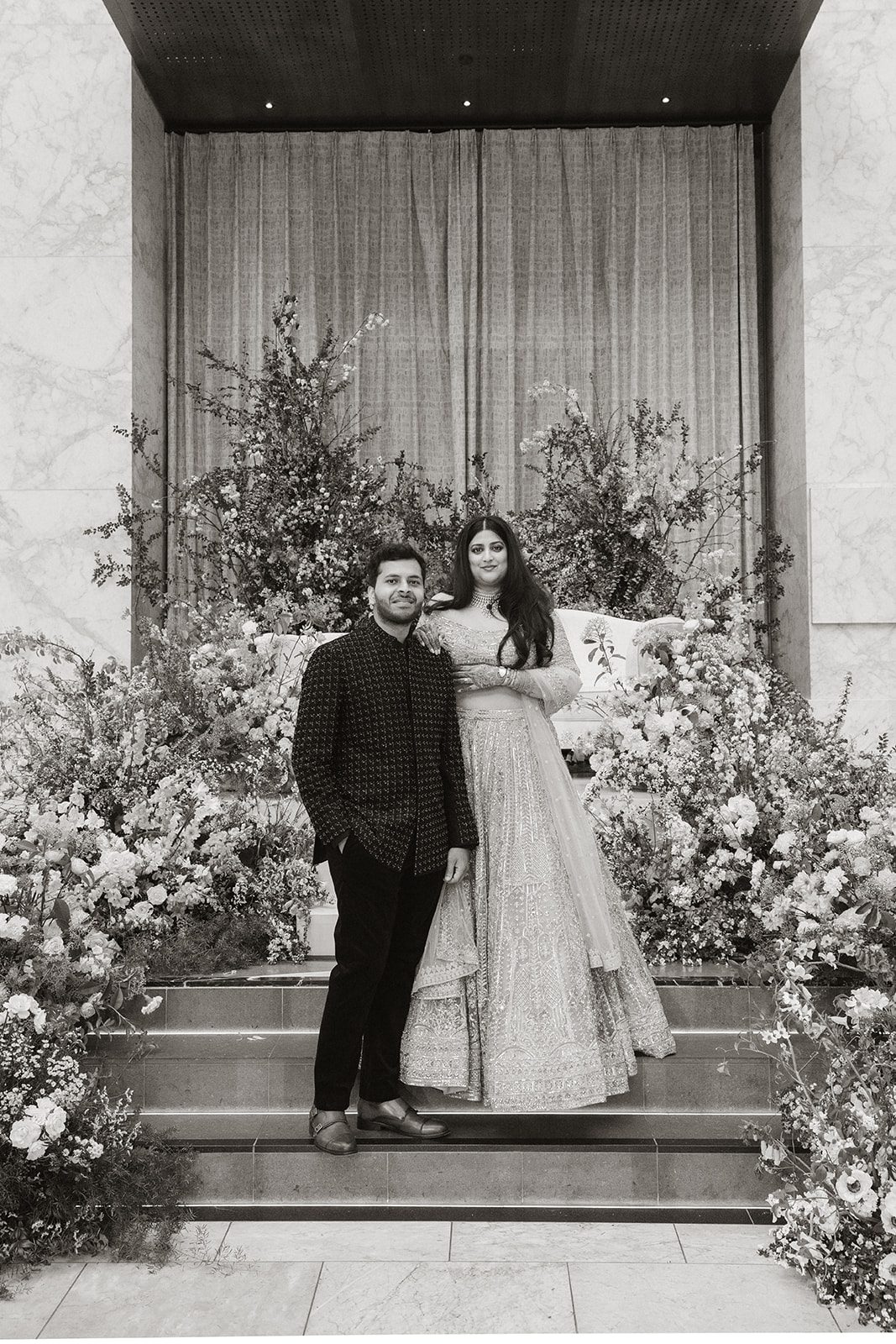 Black-and-white portrait of the couple in reception attire surrounded by floral installations during their multi-day wedding.