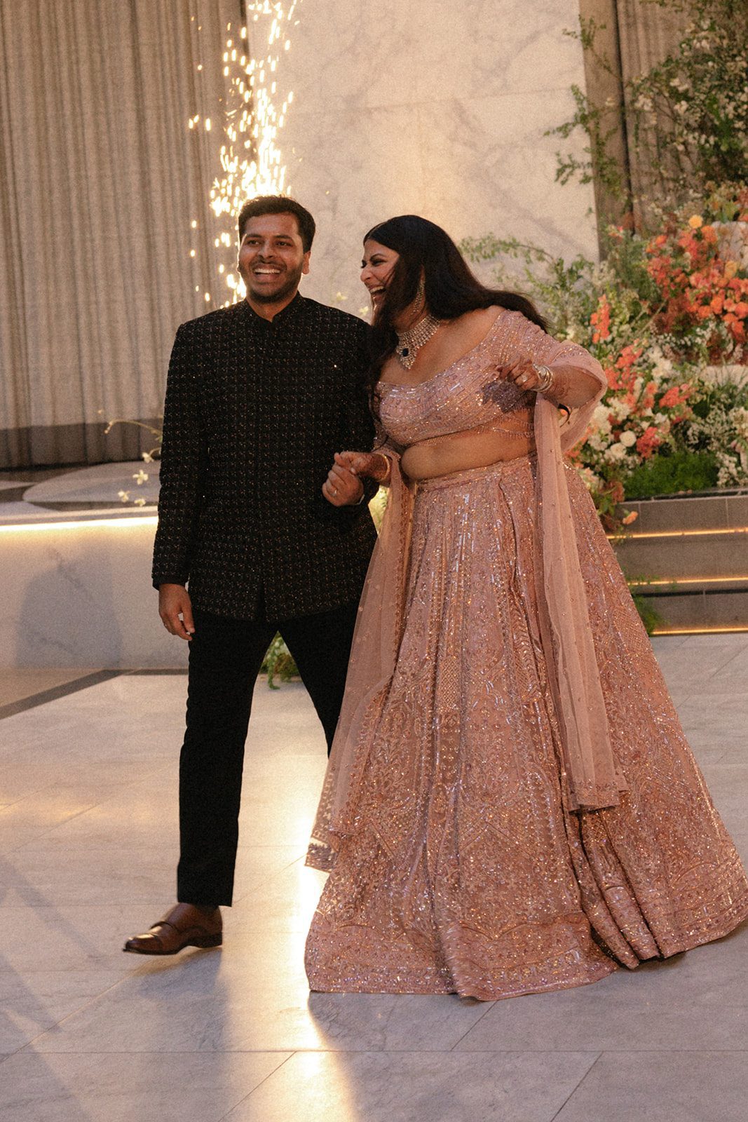 Newlyweds enter their Indian wedding reception in embellished evening attire as indoor sparklers light the celebration.