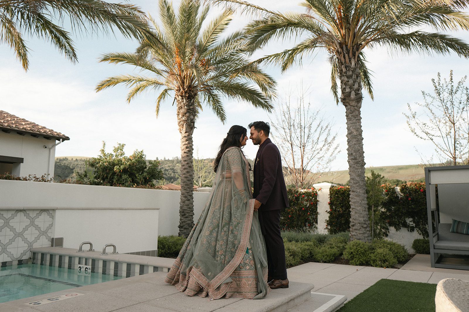Indian wedding couple standing together beneath palm trees beside a pool during their wedding celebration.