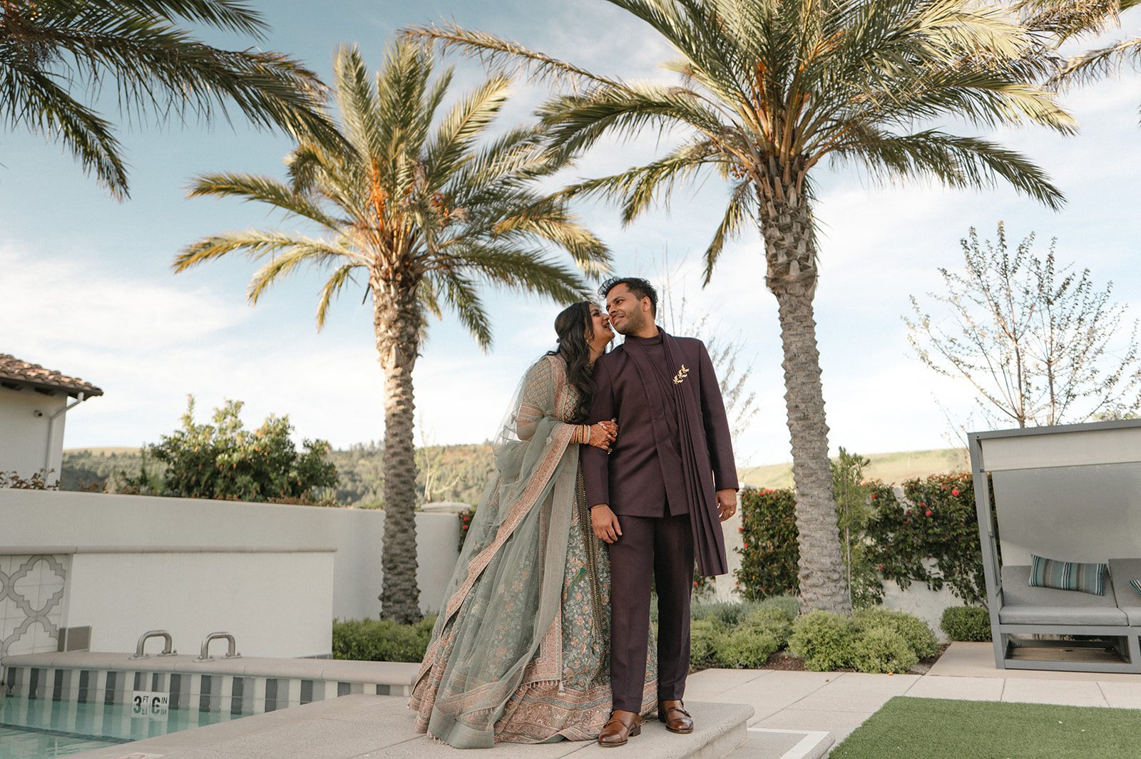 Indian wedding couple in formal attire posing beside a pool and palm trees during their multi-day wedding celebration.