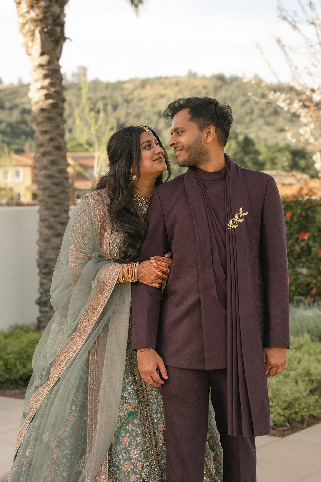 Indian wedding couple posing outdoors, with the bride in a sage green embroidered lehenga and groom in a deep plum suit.