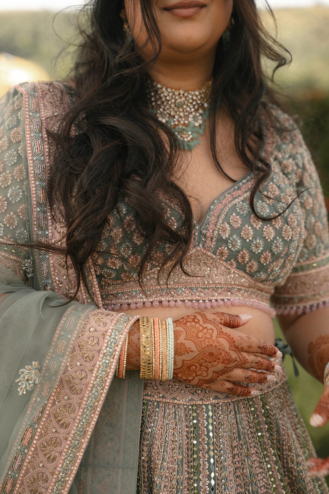 Close-up of the bride’s mehndi, bangles, jeweled necklace, and detailed sage green lehenga embroidery.