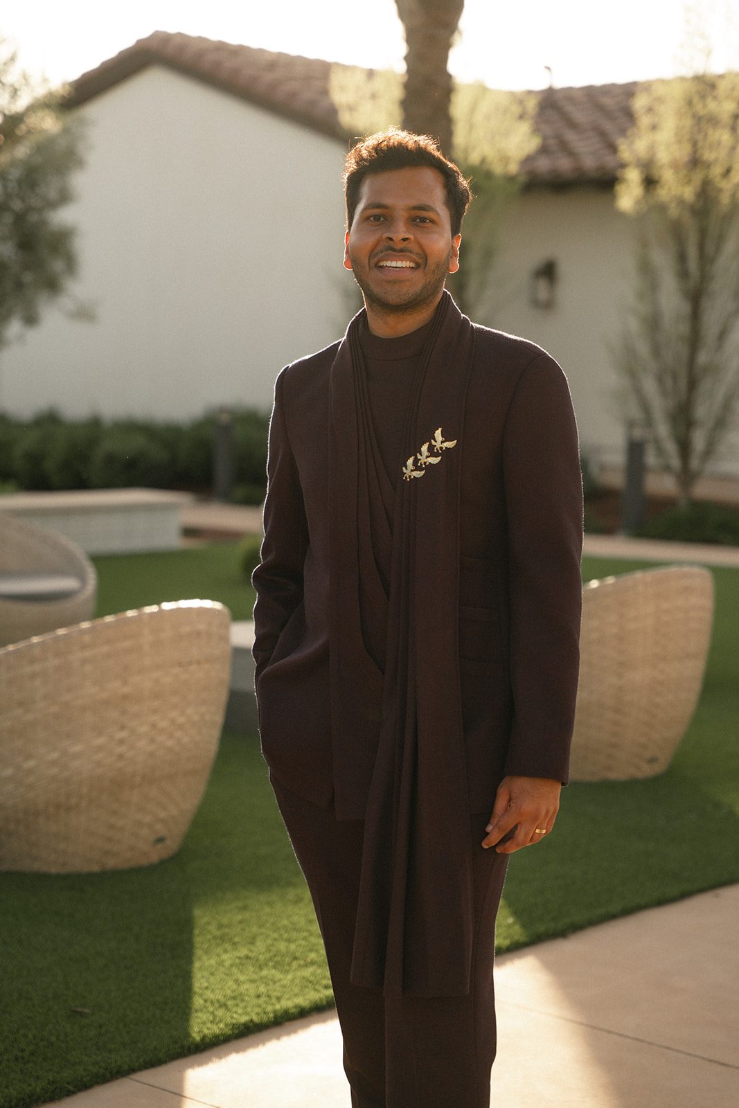 Portrait of the groom in a deep plum embroidered suit outdoors in warm evening light.