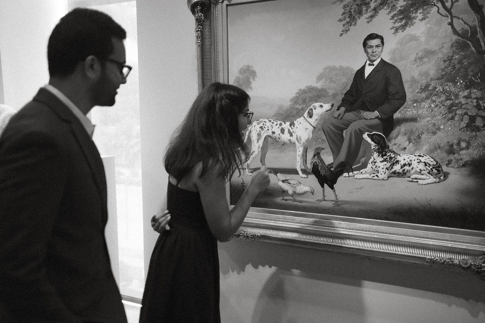 Black-and-white engagement photography of a couple examining a portrait with dogs and birds, a playful museum-inspired engagement session idea.