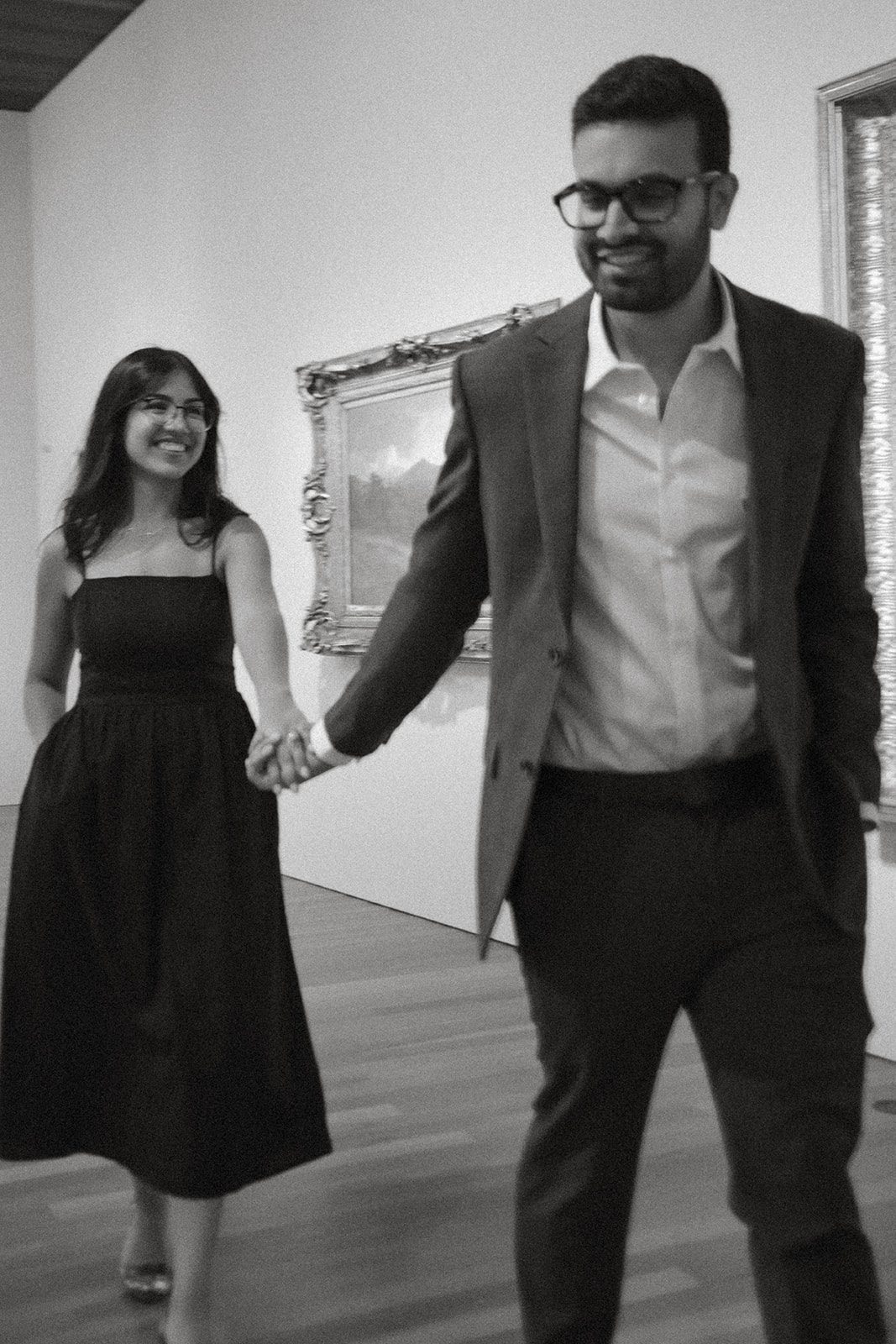 Black-and-white engagement photography of a couple smiling and holding hands while walking through a museum gallery in formal engagement session outfits.