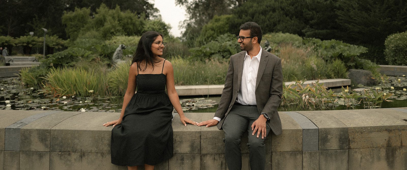 Couple sitting together beside a garden pond in coordinated formal engagement session outfits, creating a refined setting for engagement photos in San Francisco.