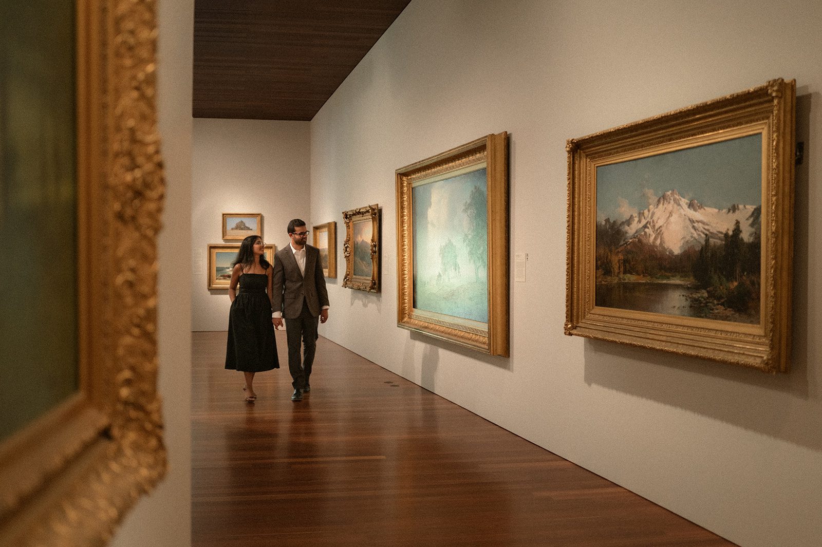 Couple holding hands while walking through a museum gallery lined with framed landscape paintings, combining elegant engagement photo outfits with an artistic setting for engagement photos in San Francisco.